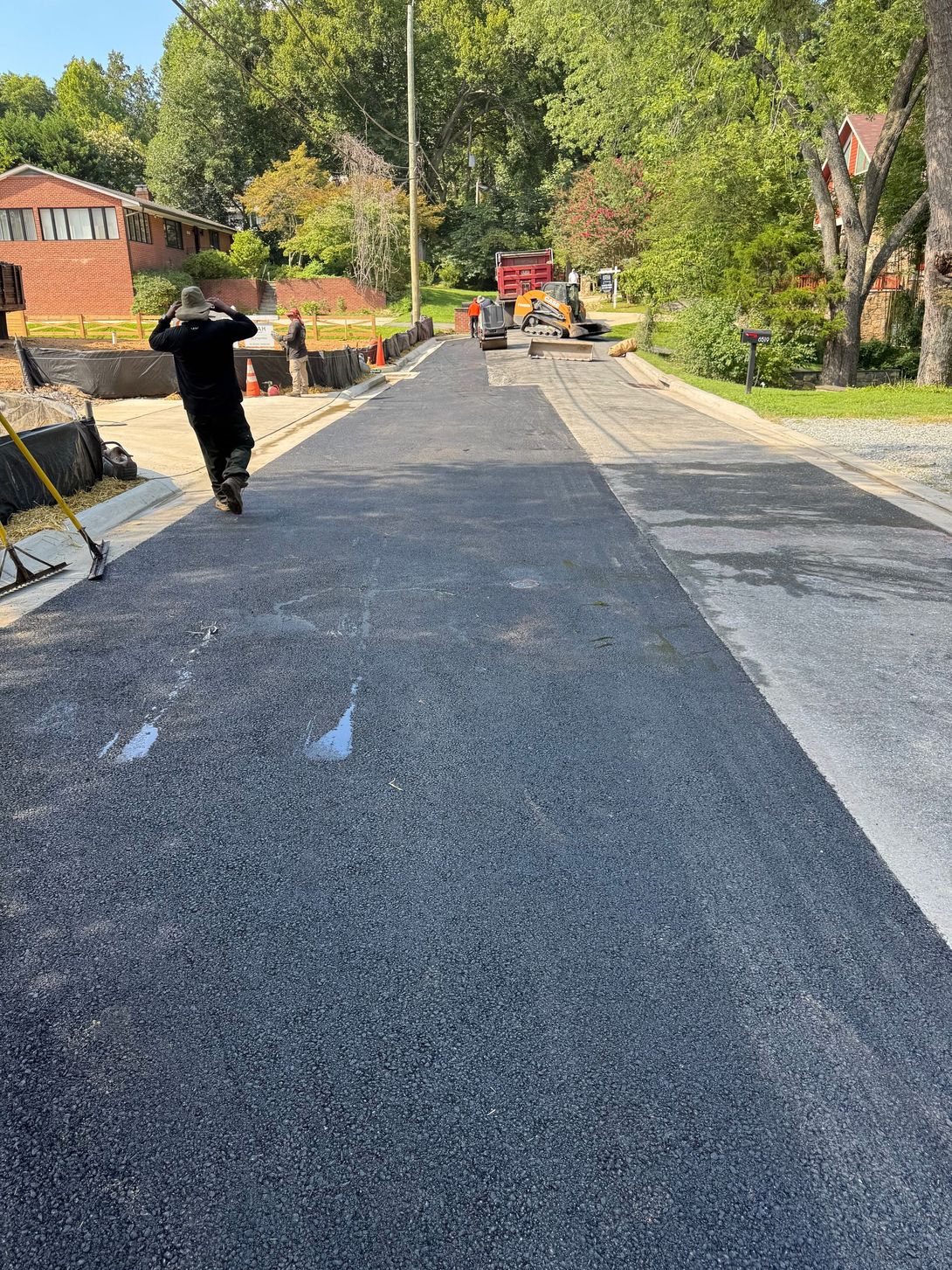 Paved road construction: Worker on scooter, fresh asphalt, cones, equipment, houses in background.