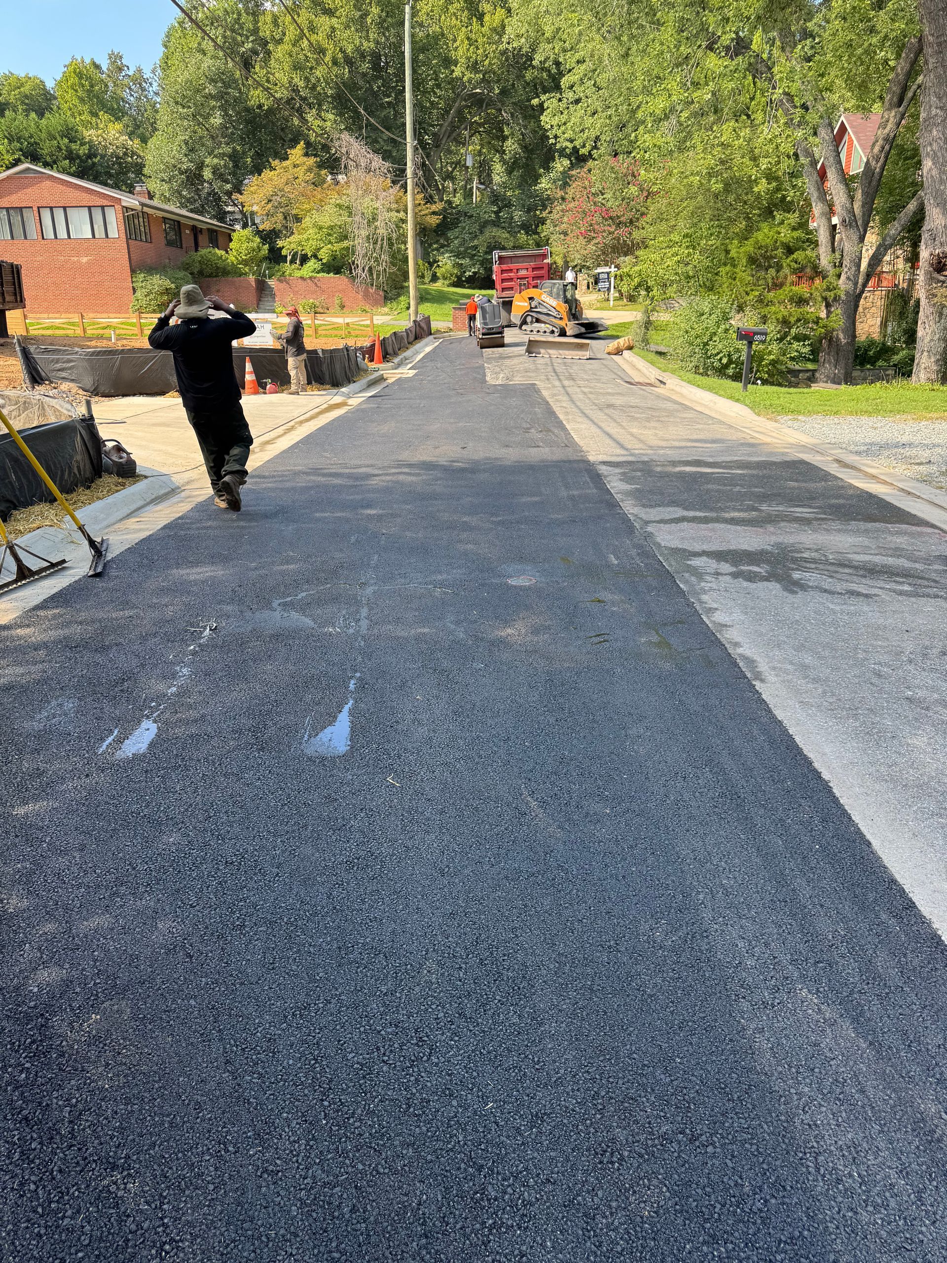 Paved road construction: Worker on scooter, fresh asphalt, cones, equipment, houses in background.