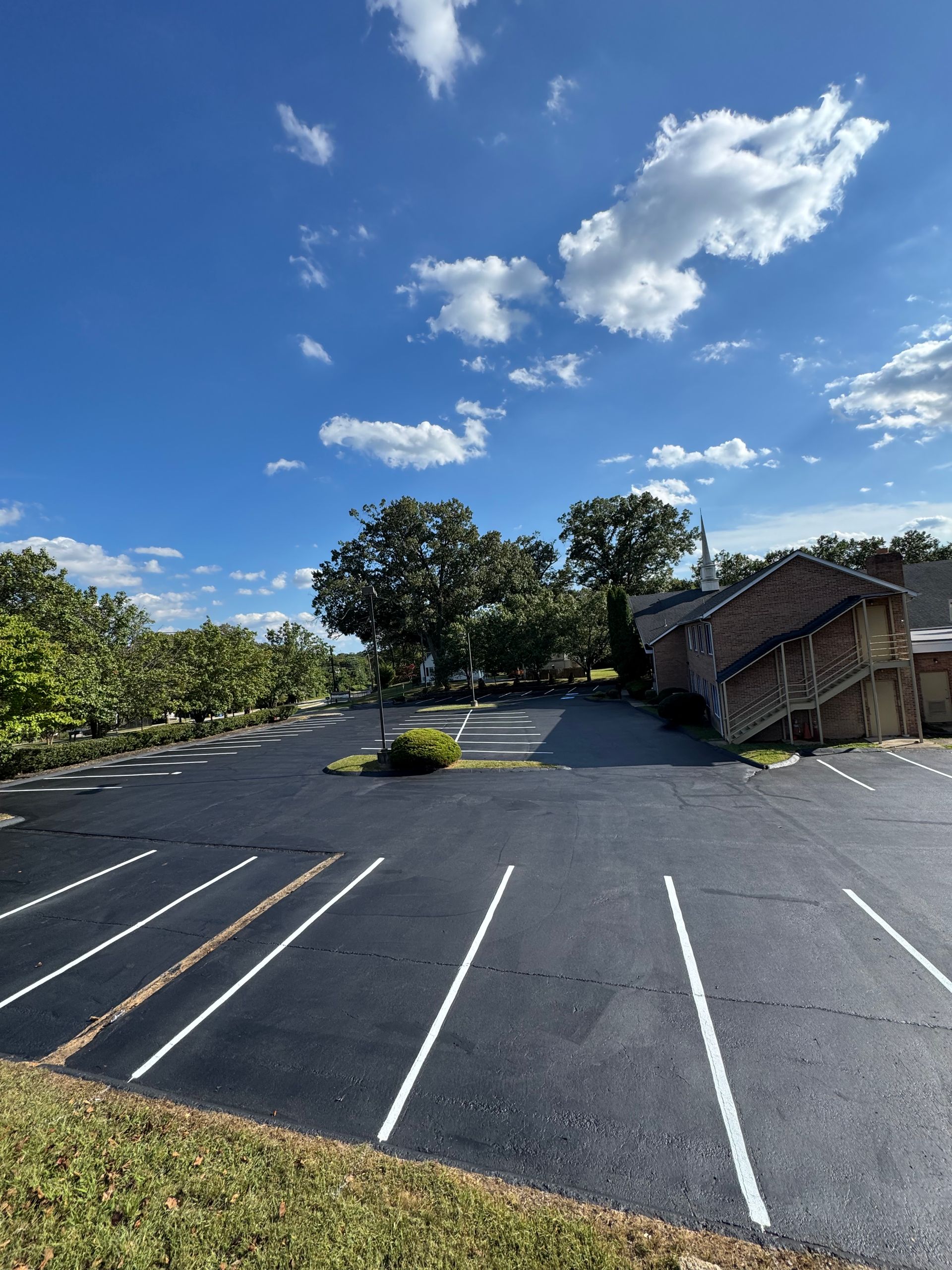 Empty parking lot with white lines, brick building, trees under blue sky.