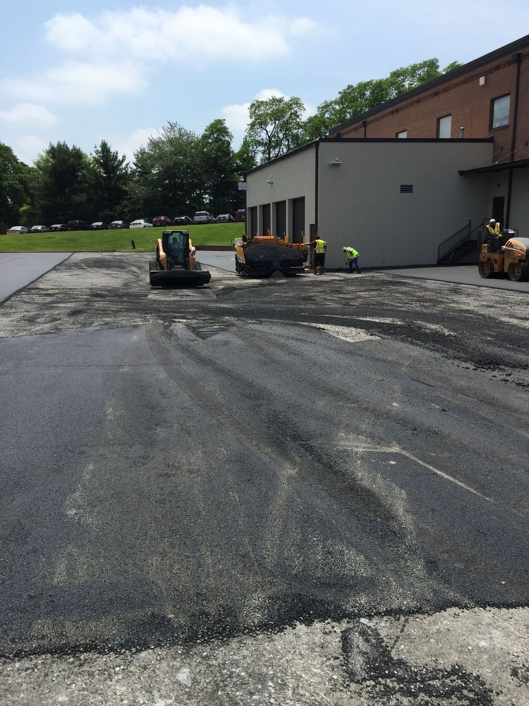 Workers paving a parking lot next to a building. Asphalt is being spread by machinery on a sunny day.