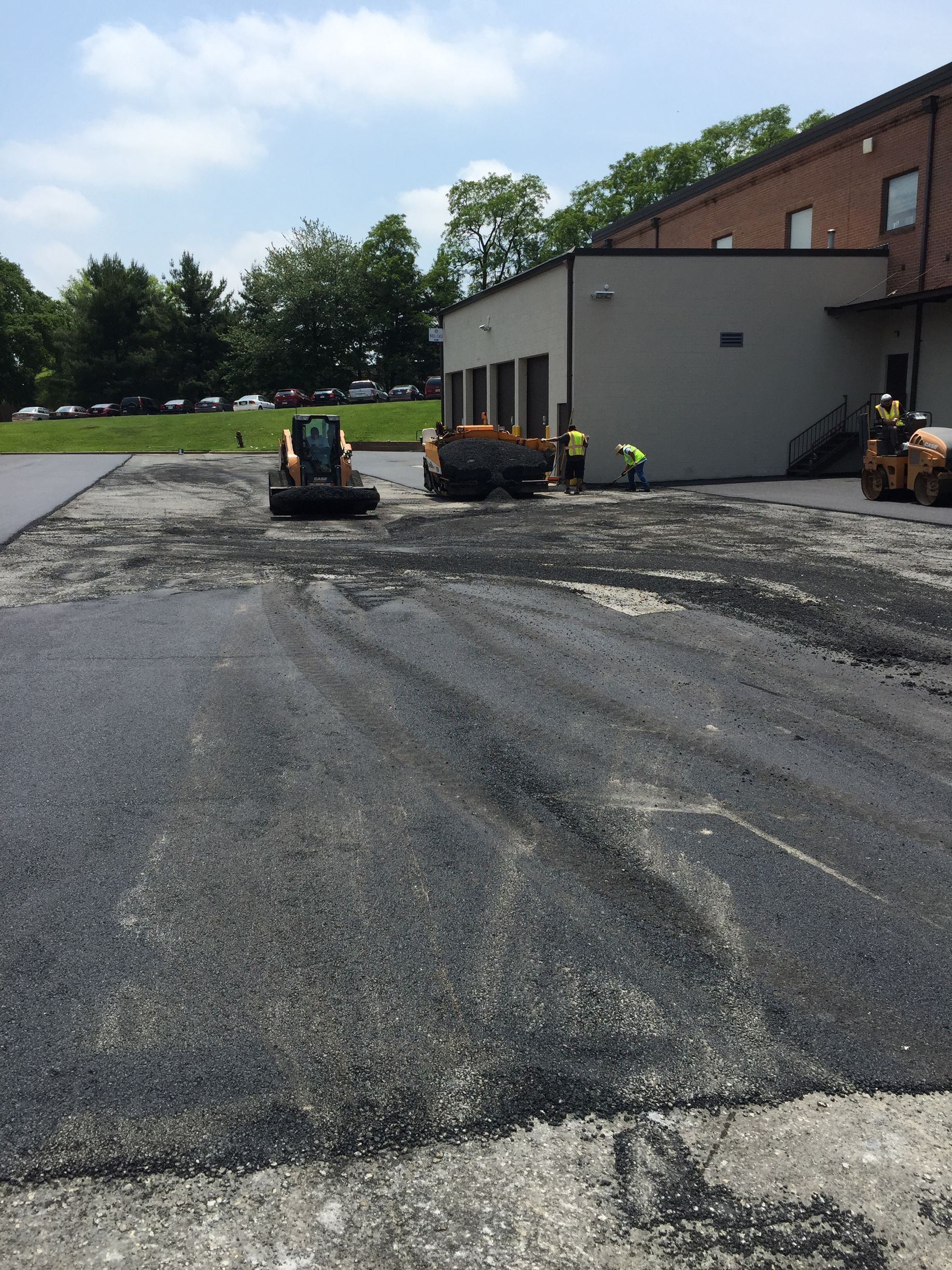 Workers paving a parking lot next to a building. Asphalt is being spread by machinery on a sunny day.