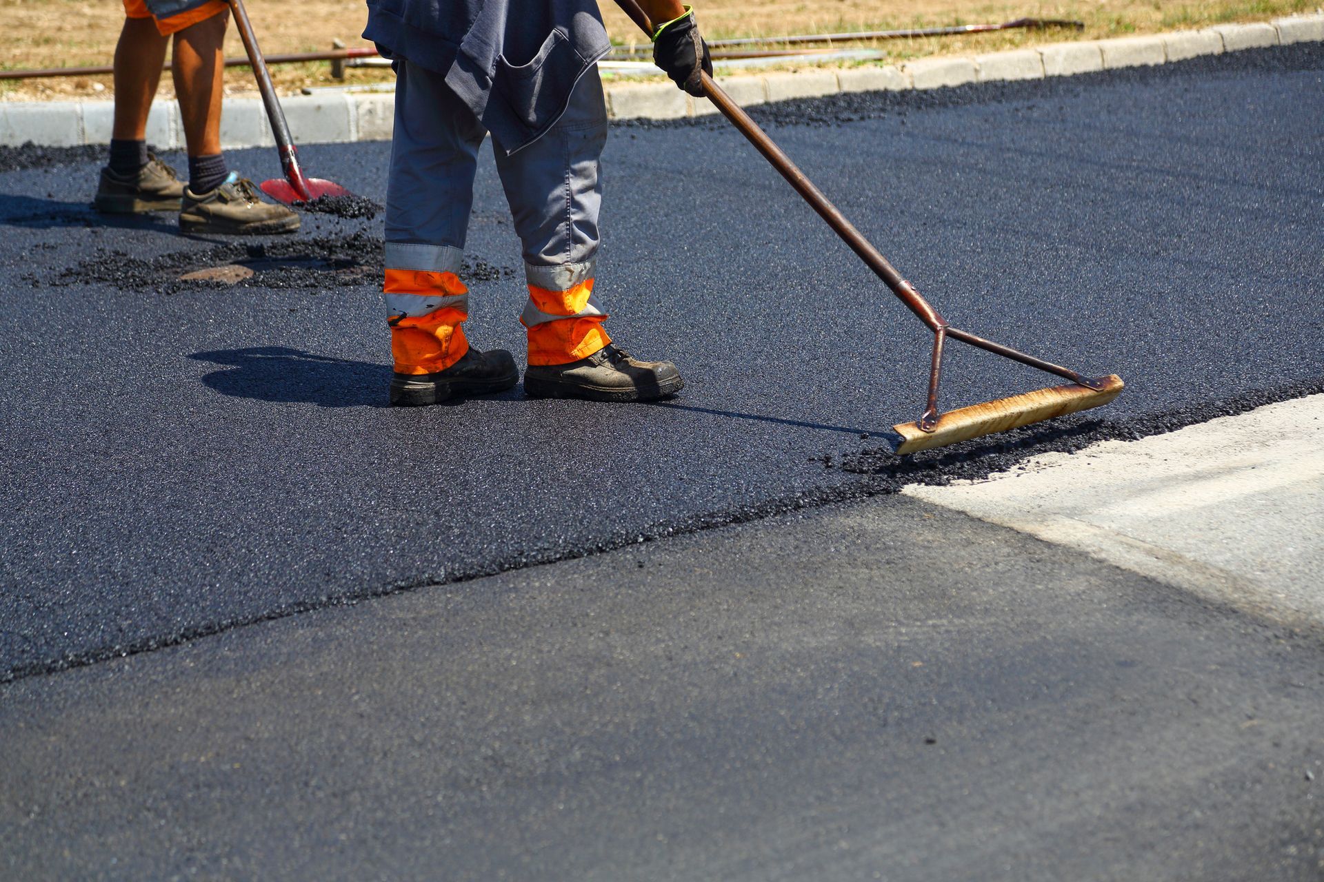 Workers paving a road, using long-handled rakes to smooth the fresh asphalt.