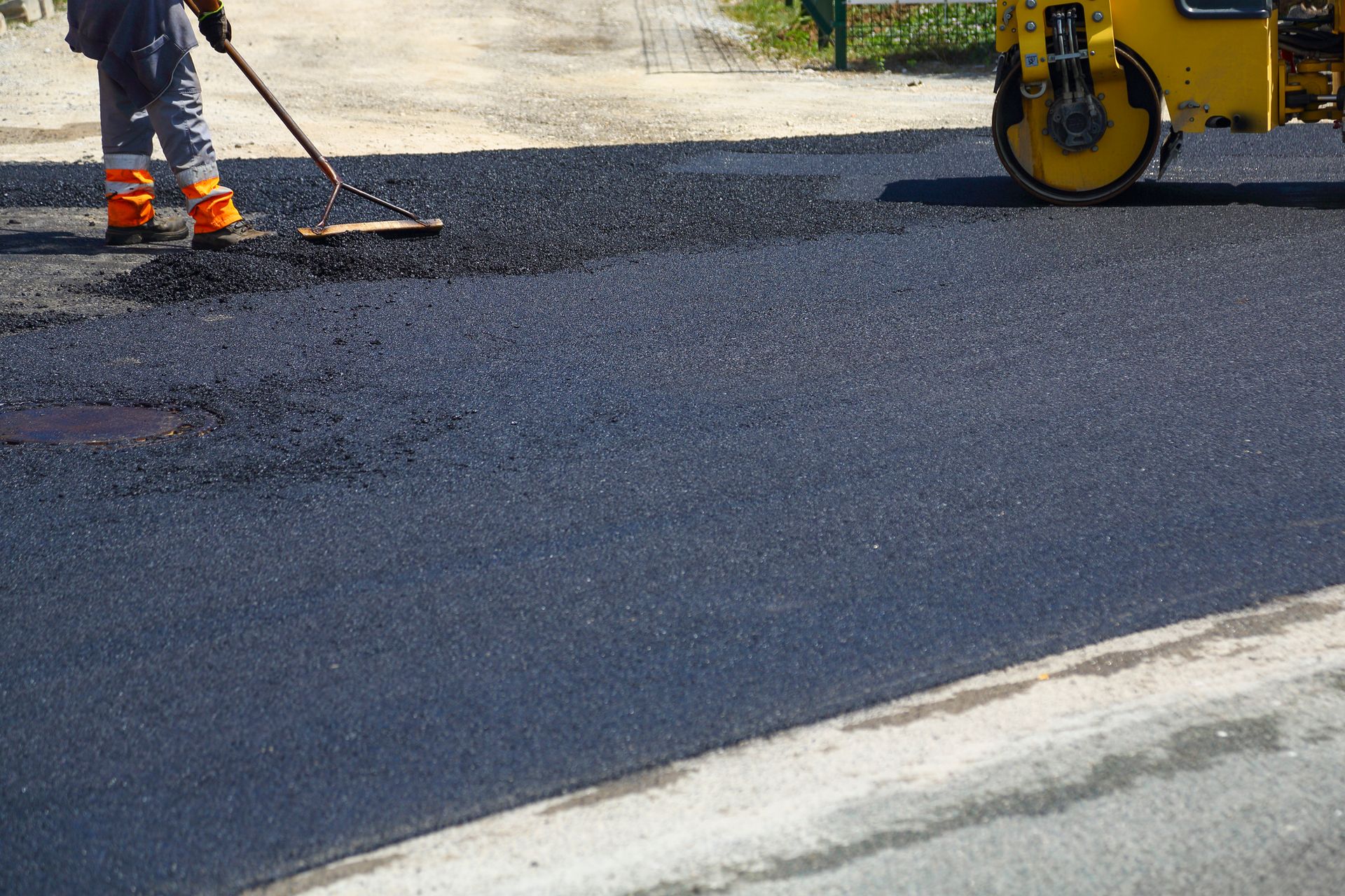 Road construction: worker using a rake to smooth fresh asphalt. Yellow roller compacting the surface.