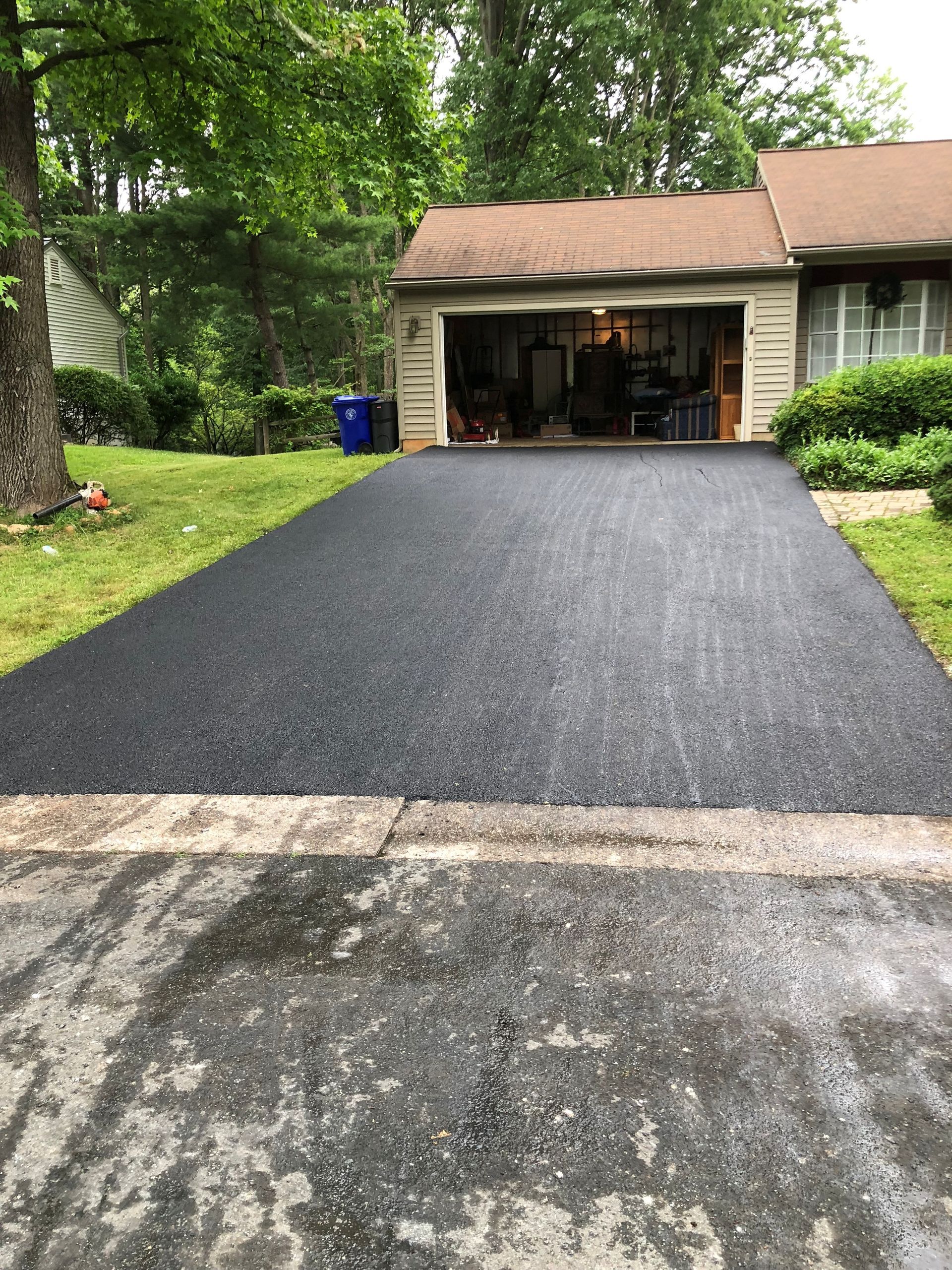 A newly paved, dark asphalt driveway leads to an open residential garage on a sunny day with trees in the background.