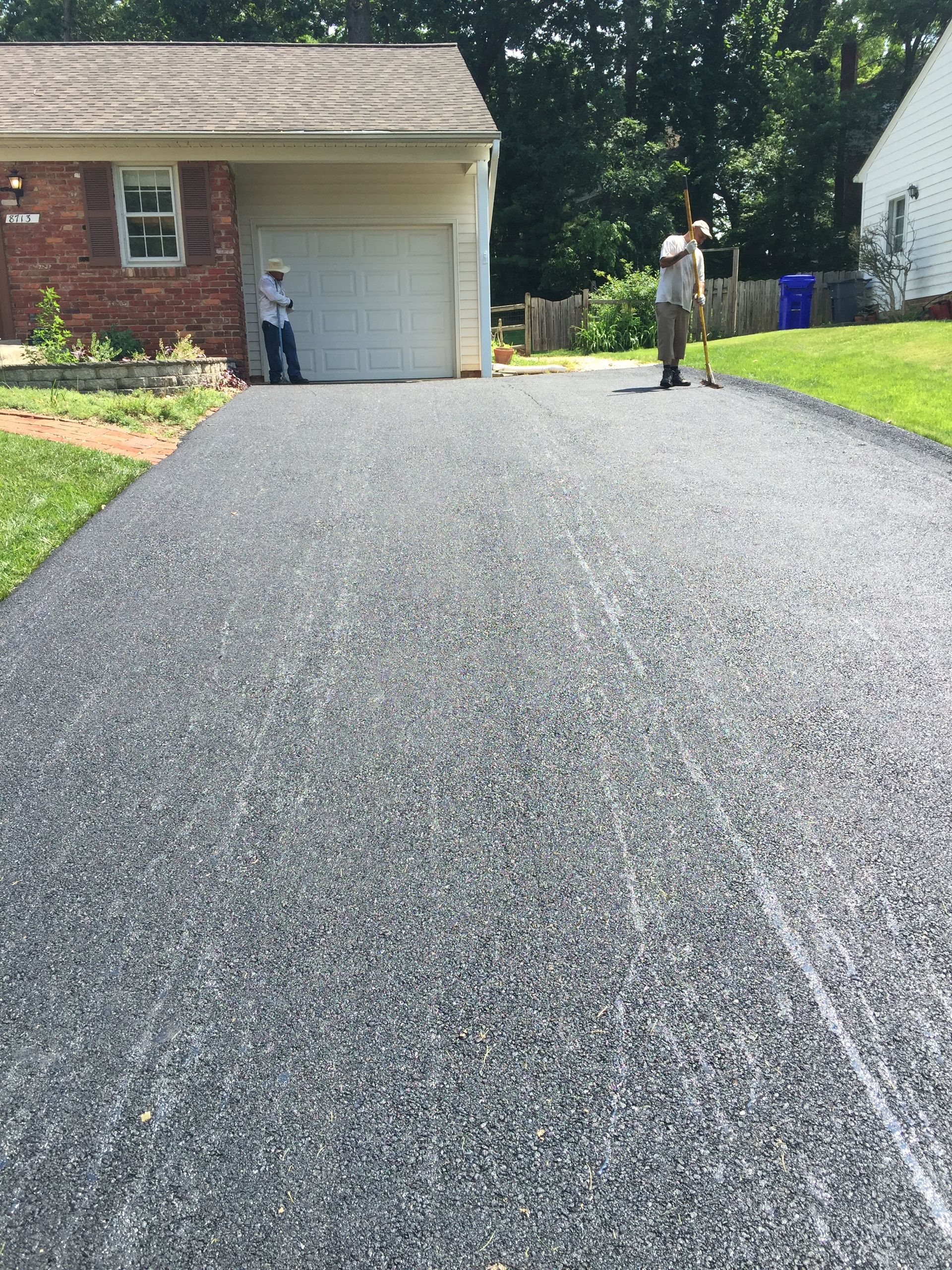 Two people stand by a brick house and a newly paved asphalt driveway on a sunny day.