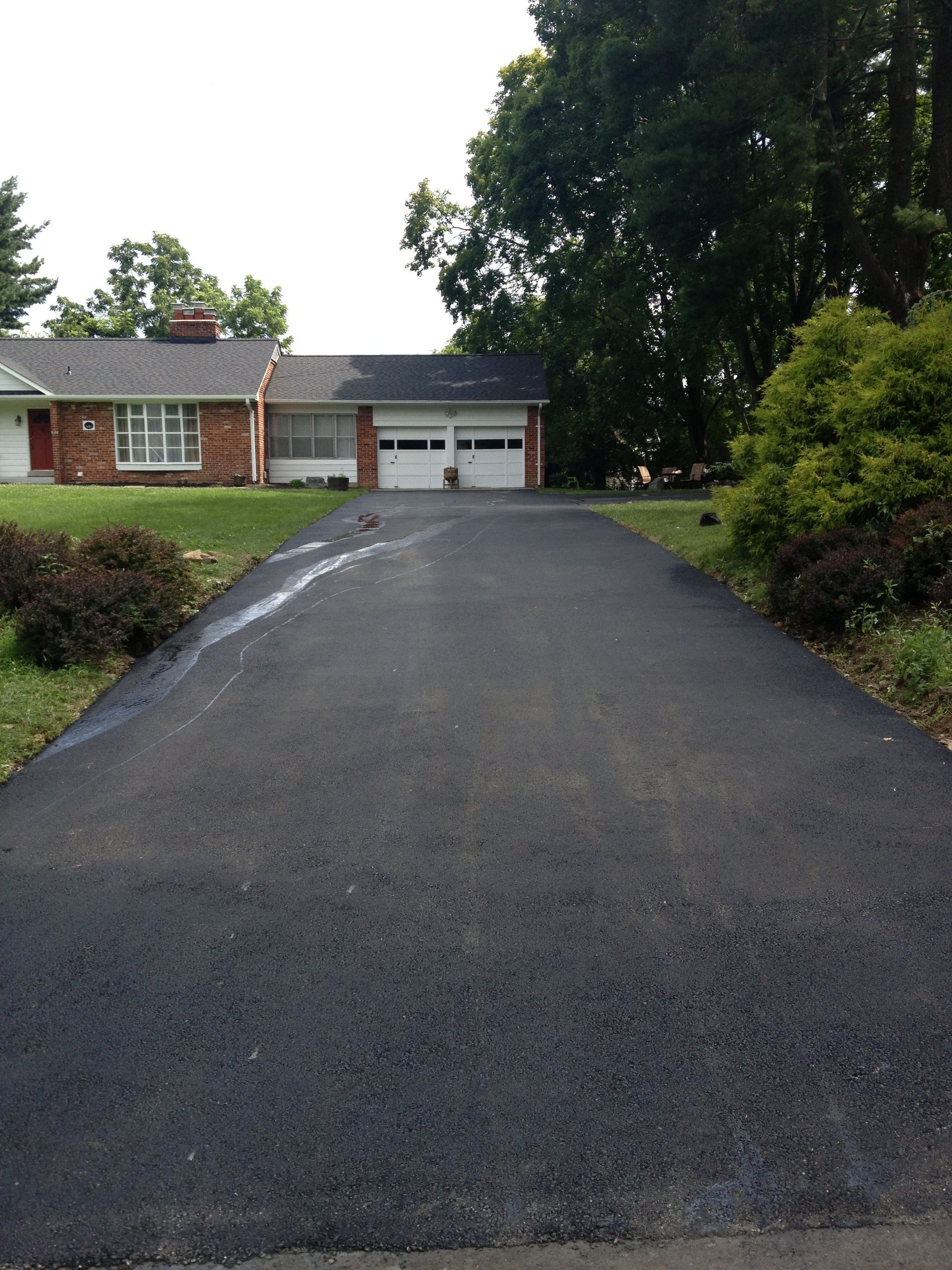 A newly paved asphalt driveway leads to a brick house with an attached two-car garage under a bright sky.