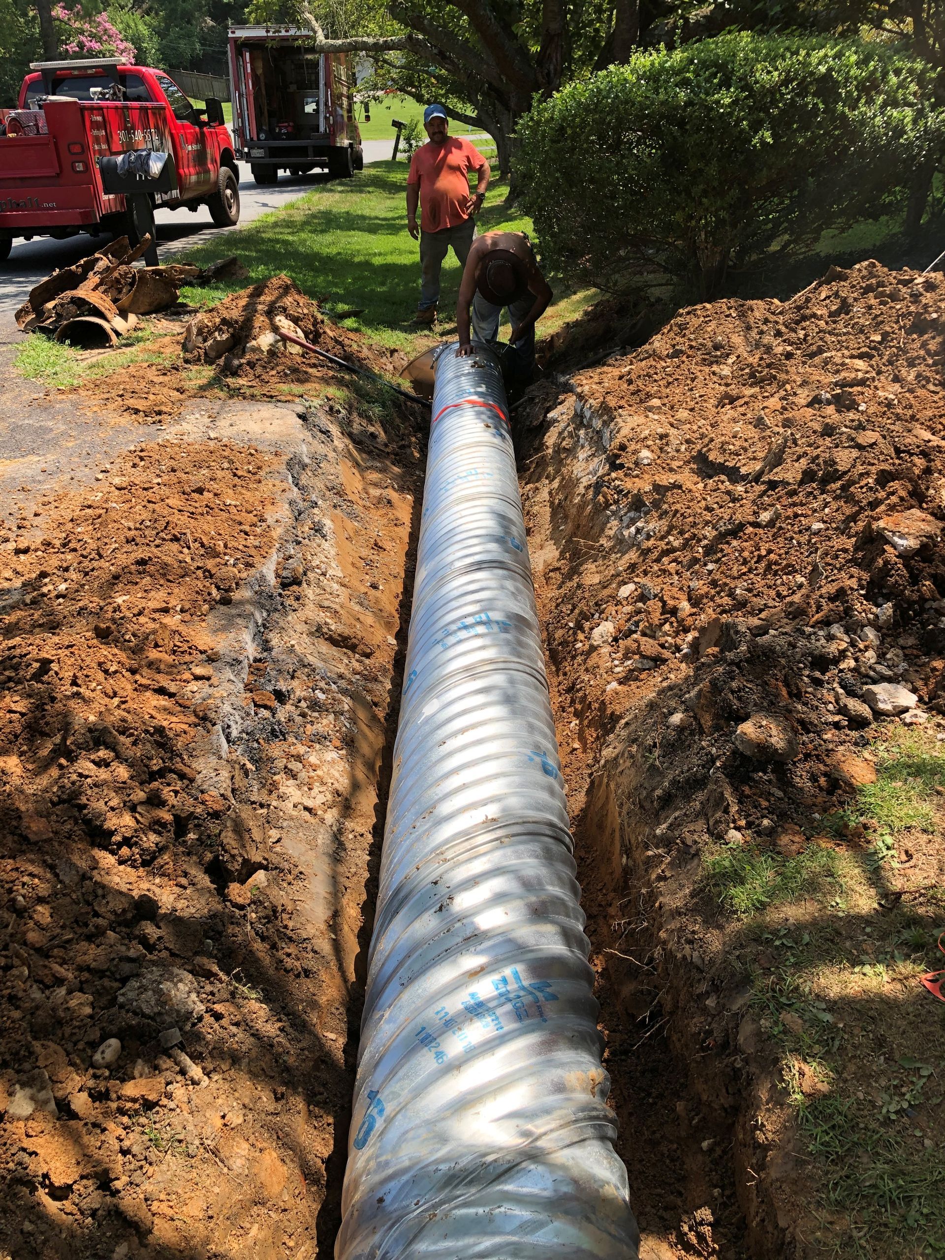 Two workers installing a large, corrugated metal pipe in a narrow dirt trench with utility trucks parked nearby.