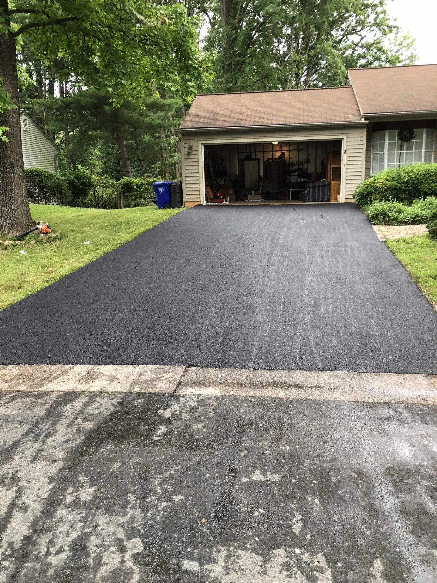 Freshly paved asphalt driveway leading to a garage; green lawn and trees in the background.