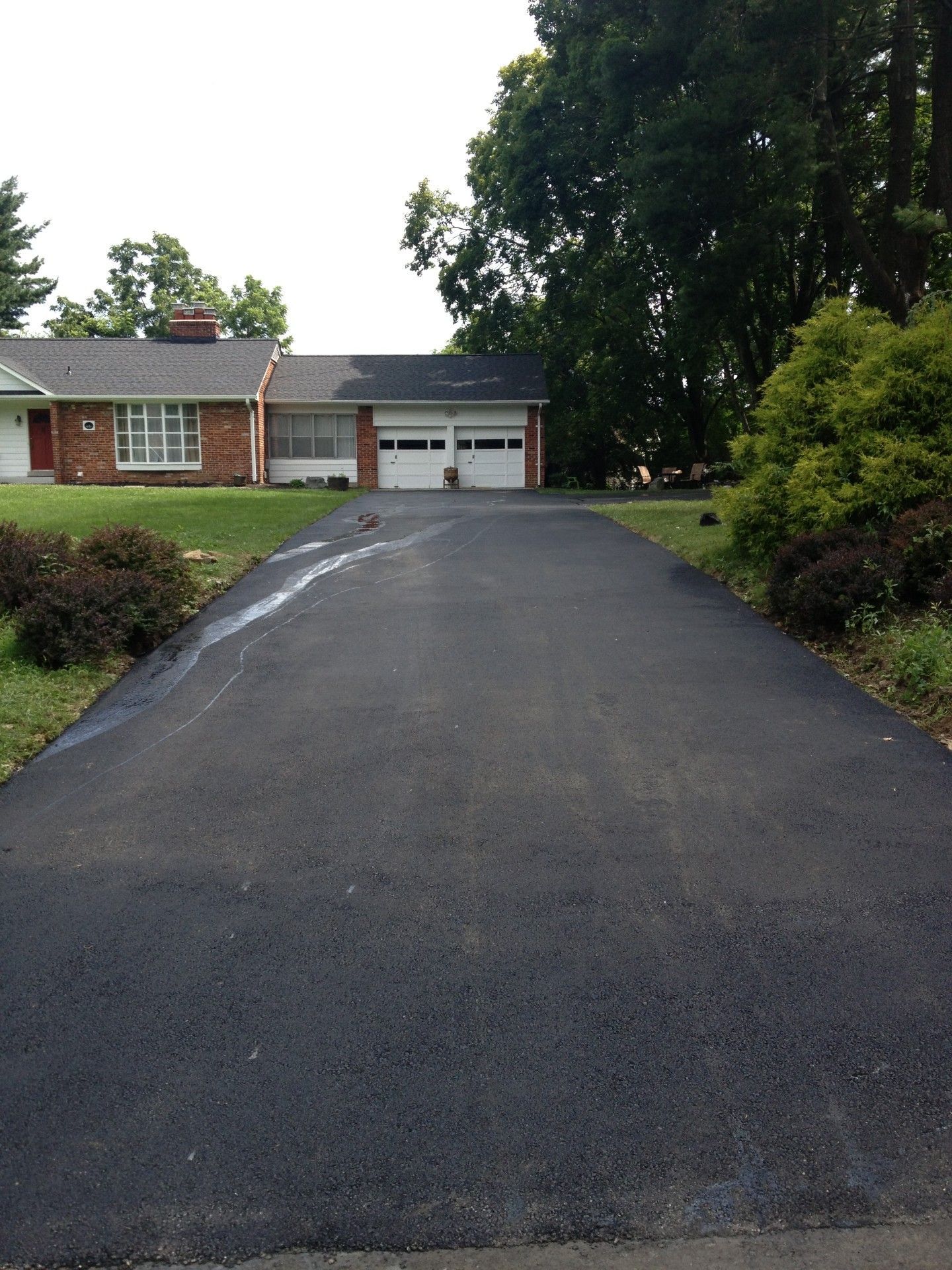 Black asphalt driveway leads to a brick house with a two-car garage; overcast sky.