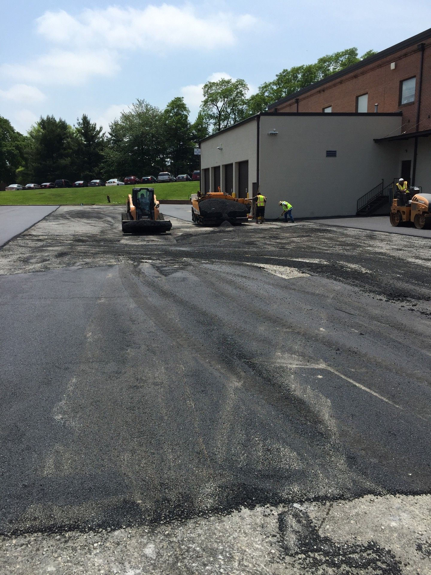 Asphalt paving in progress: workers, machinery, dark asphalt surface, near a building with trees and blue sky.