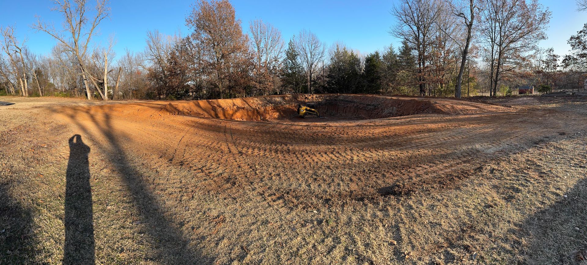 A barren landscape with a dirt pit in the foreground and trees under a blue sky in the background.