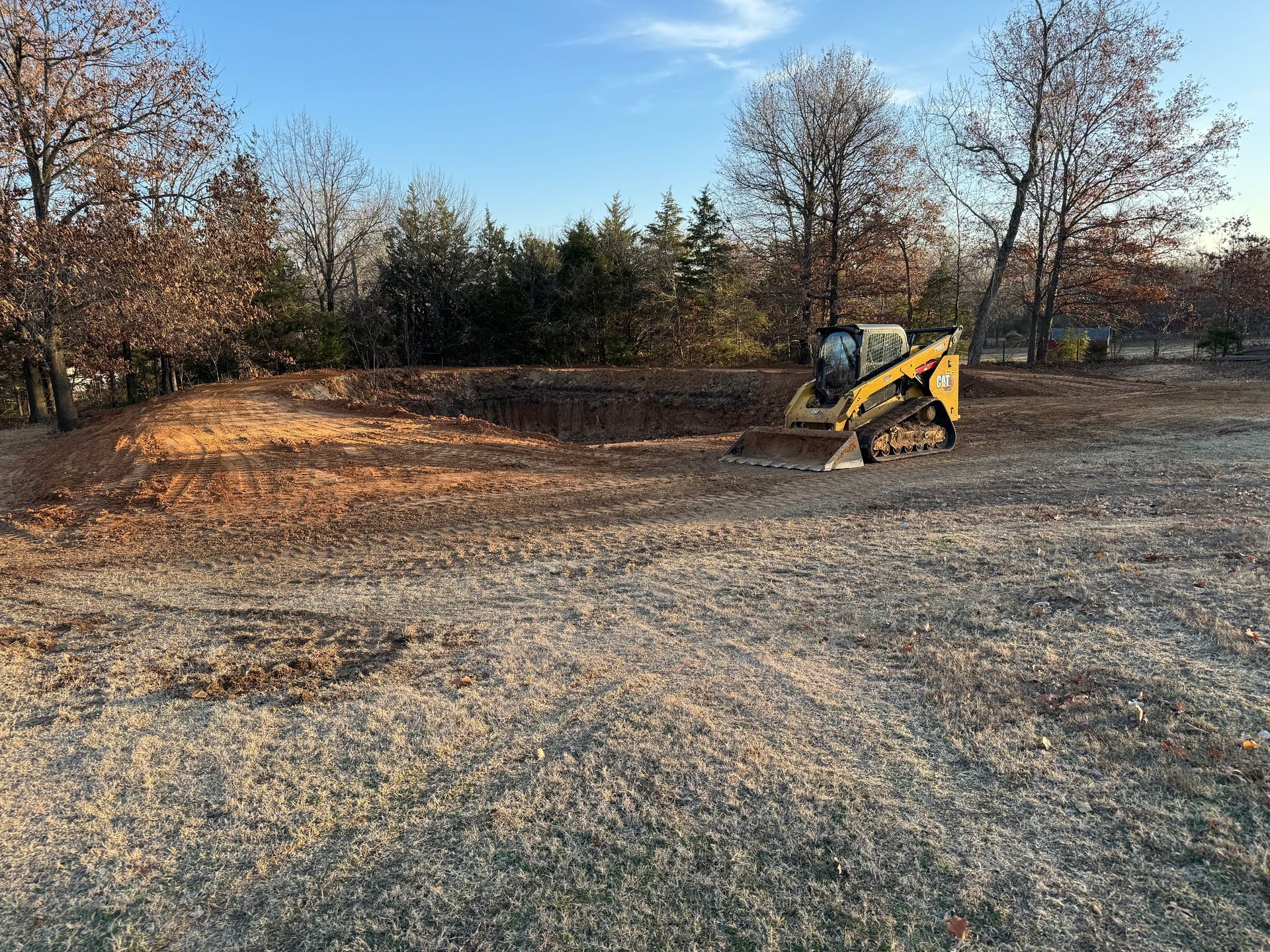 A compact track loader on a dirt pile, in a clearing near trees on a sunny day.