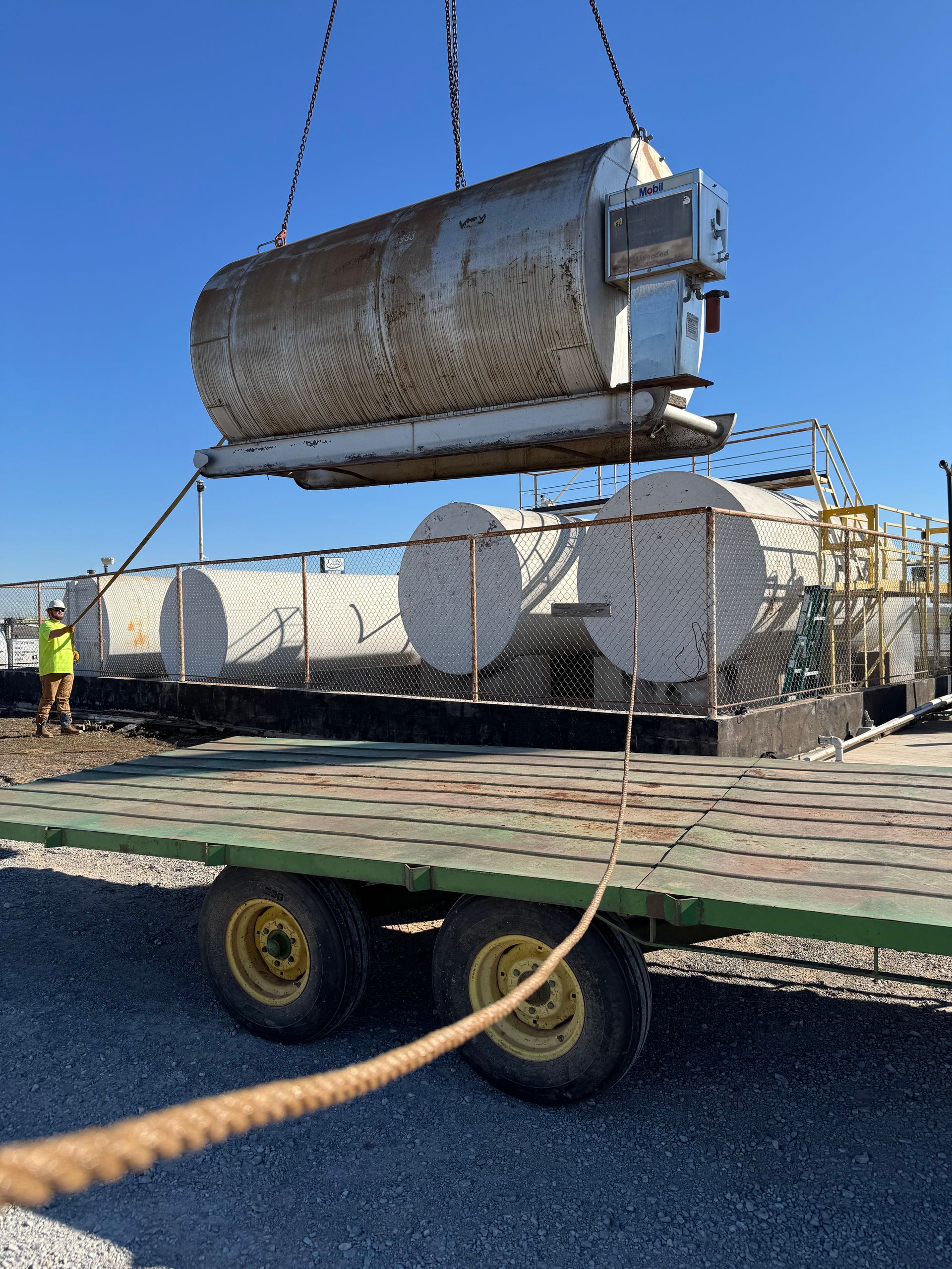 A large tank being lifted by a crane onto a flatbed trailer, with two other tanks nearby. Blue sky.