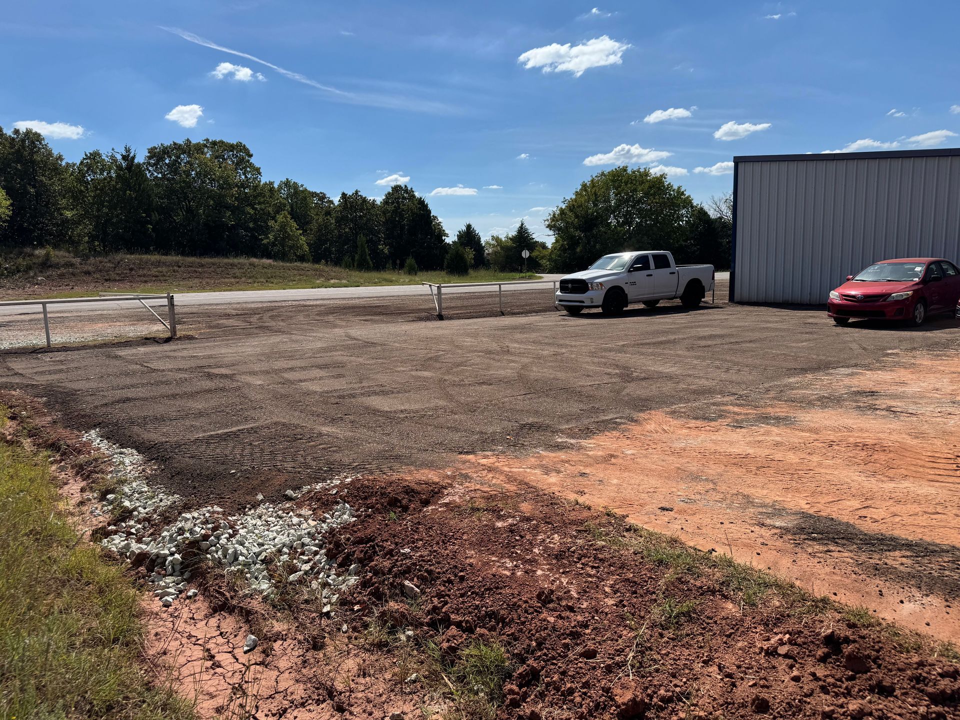Gravel parking area next to a metal building, with a white truck and red car parked.