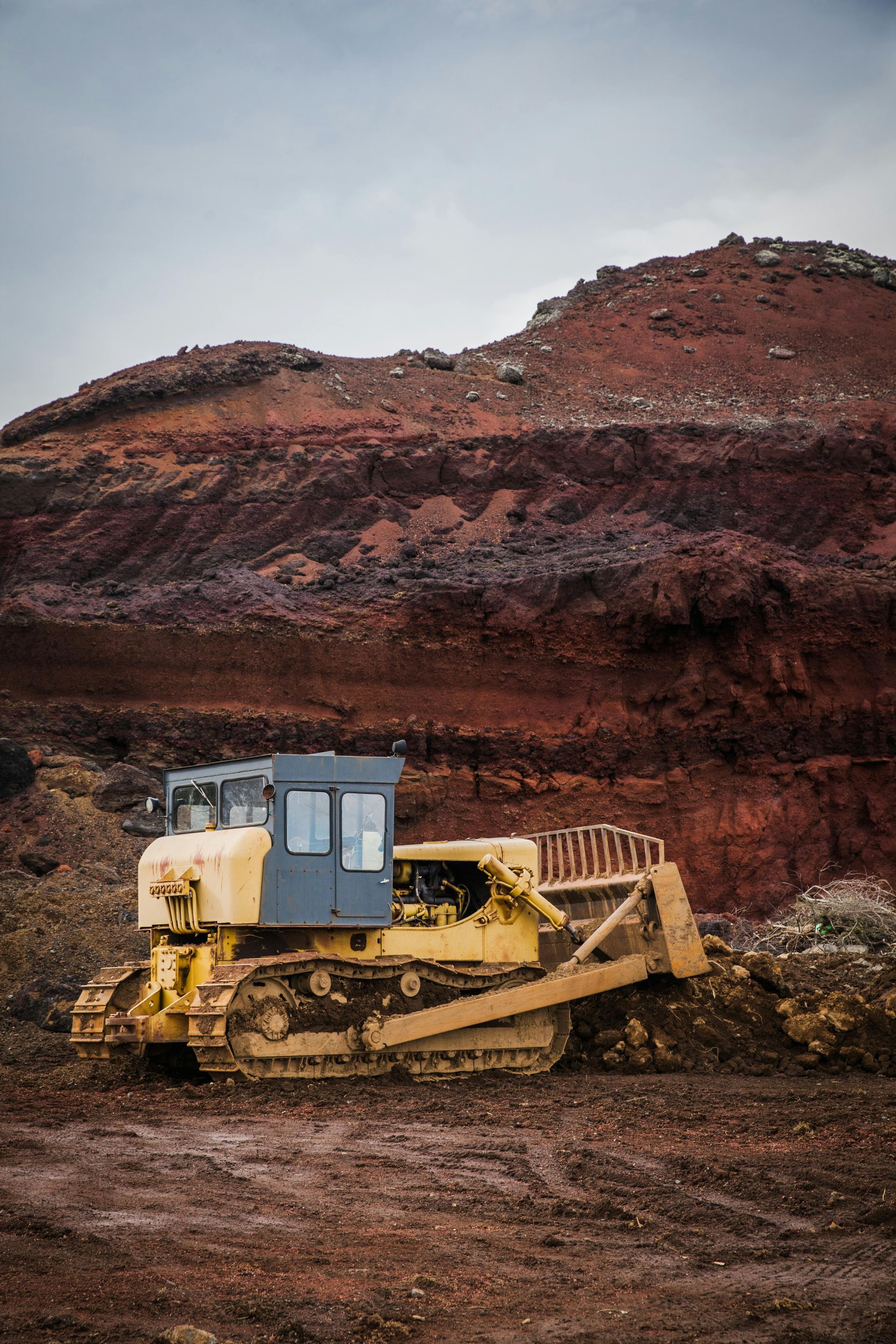 Yellow bulldozer clearing earth in front of a reddish-brown rock formation. Cloudy sky.