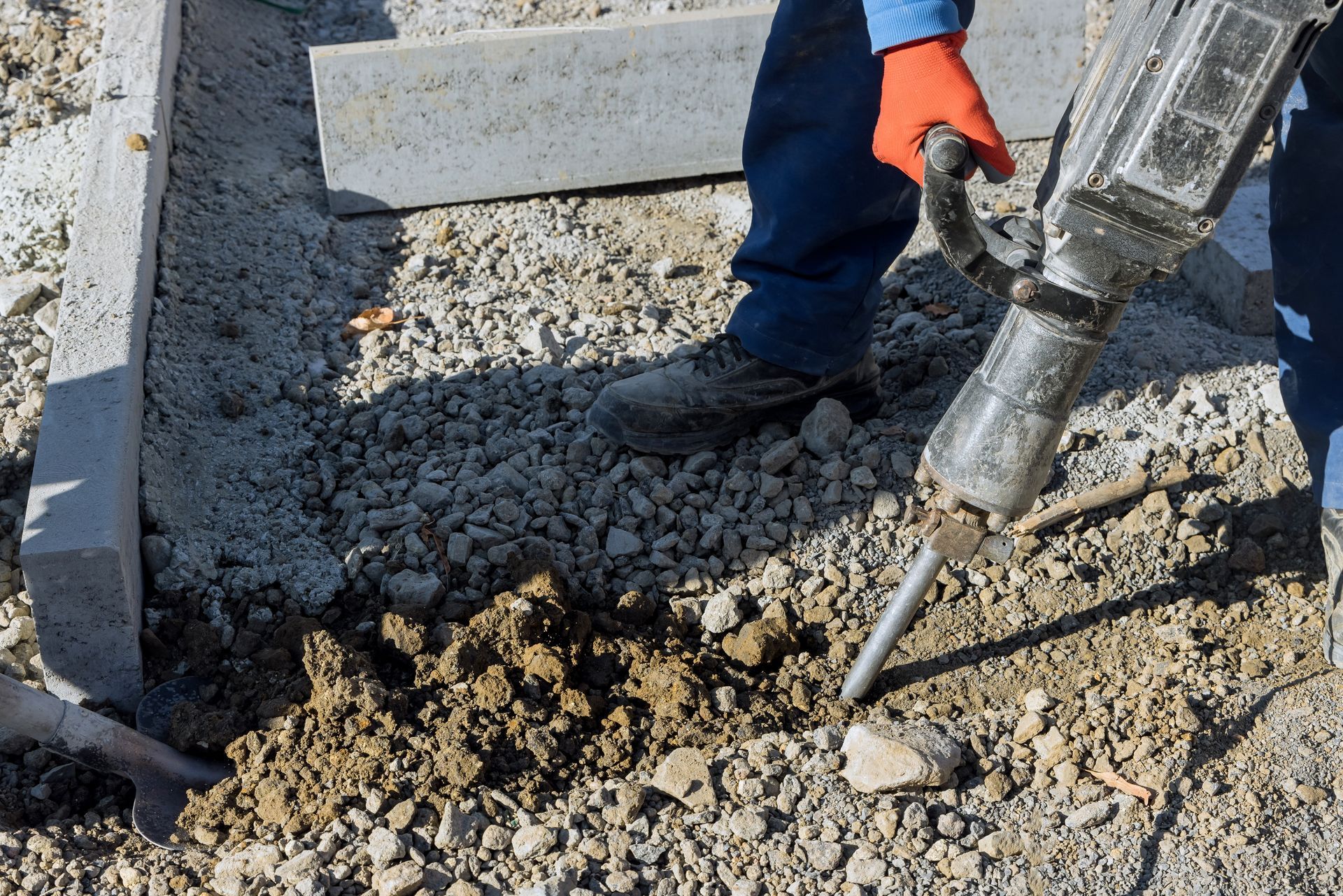Person in blue work clothes using a jackhammer on dirt and gravel near concrete blocks.