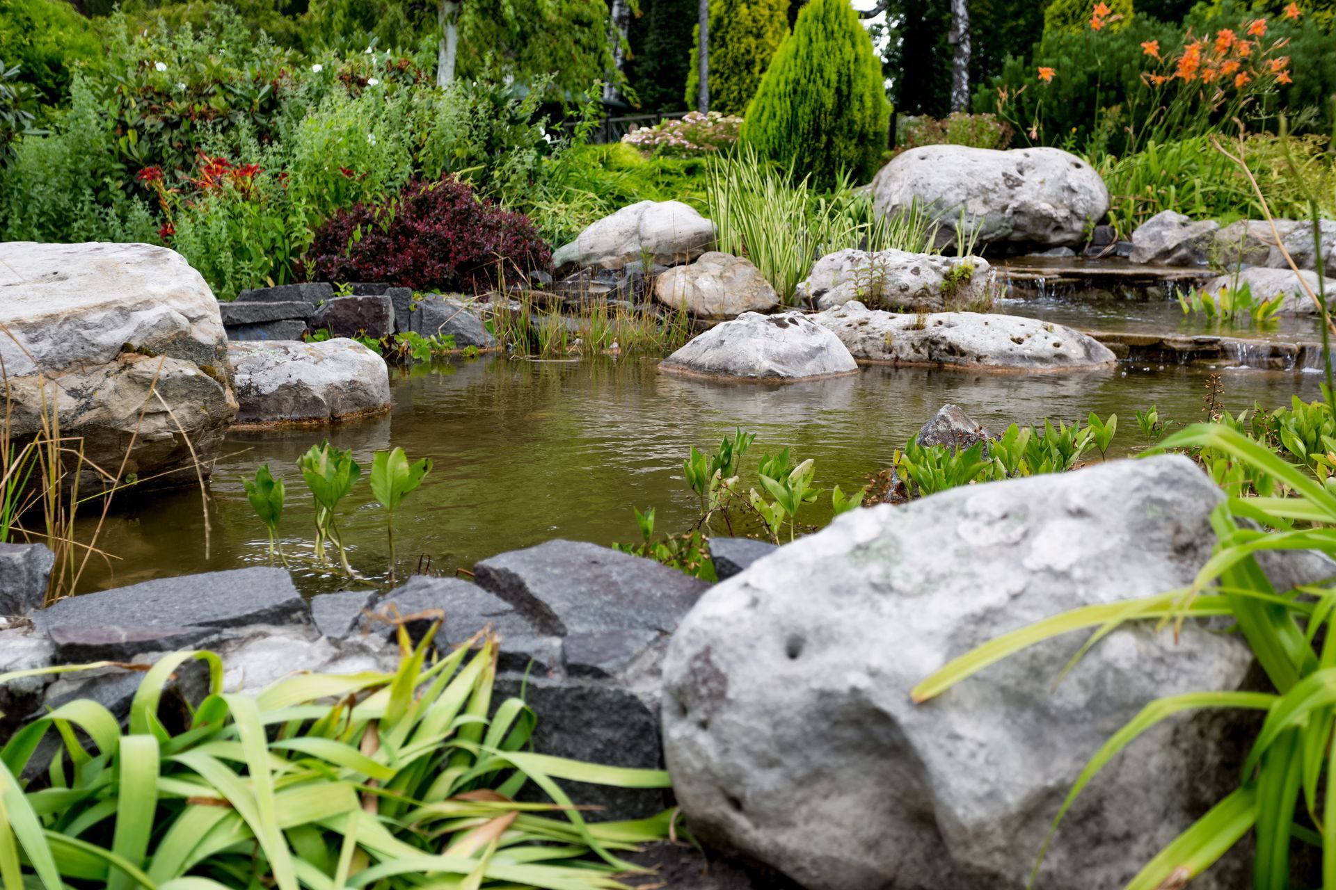 A tranquil garden pond with large rocks, greenery, and small cascading water features.