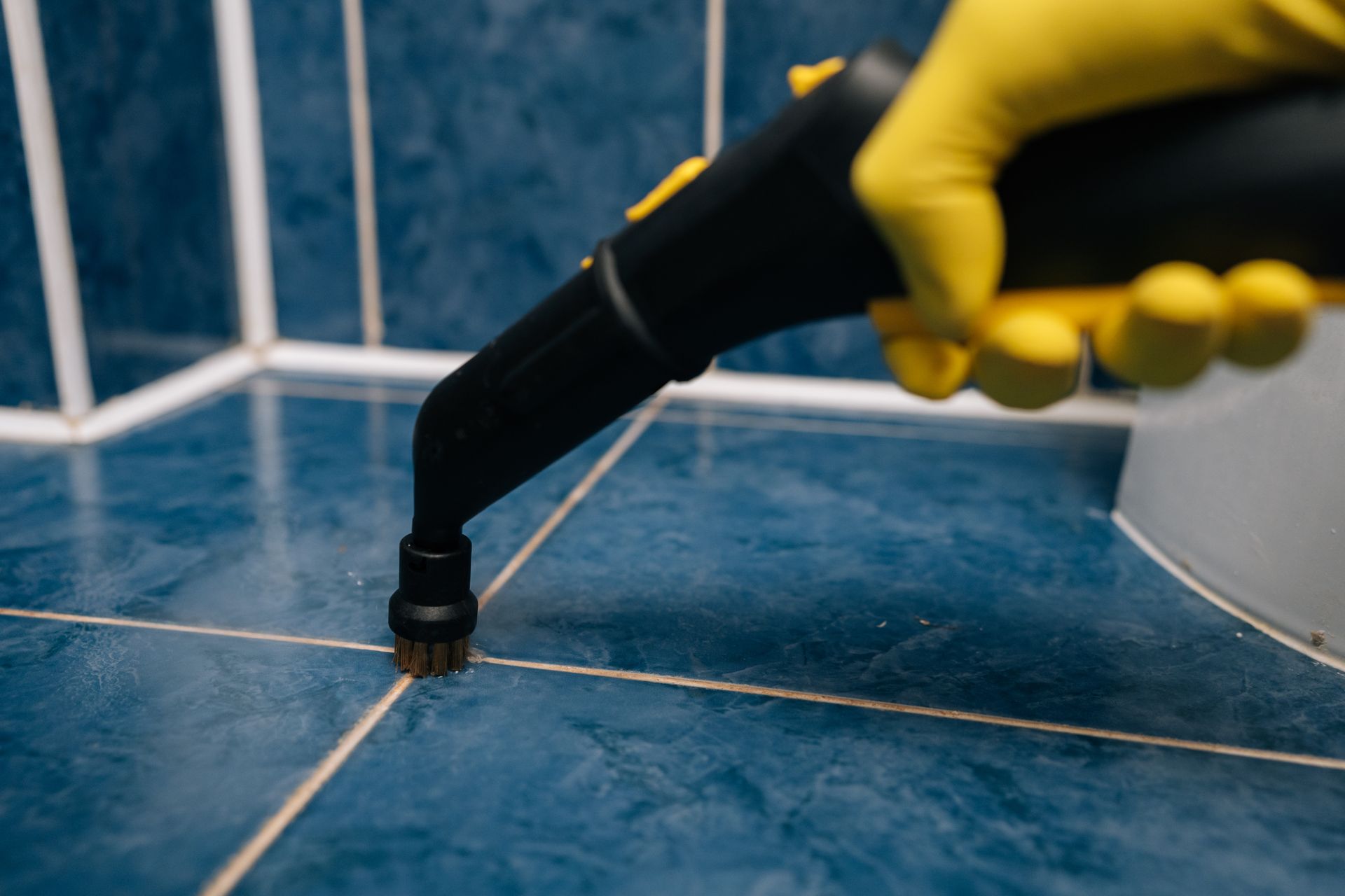 Yellow-gloved hand using a steam cleaner nozzle on blue tile grout at a shower corner Yellow-gloved hand using a steam cleaner nozzle on blue tile grout at a shower corner