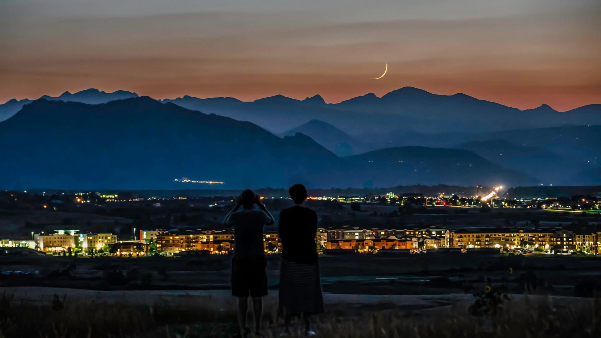Two people overlook a lit city at dusk, with mountains and a crescent moon in the background.