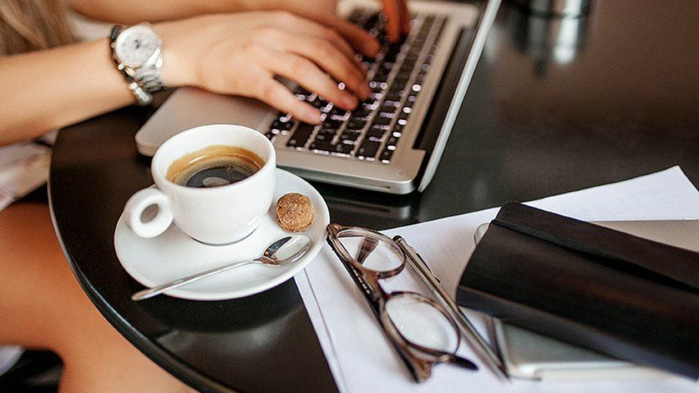 Hands typing on a laptop beside coffee, glasses, and a notebook on a desk