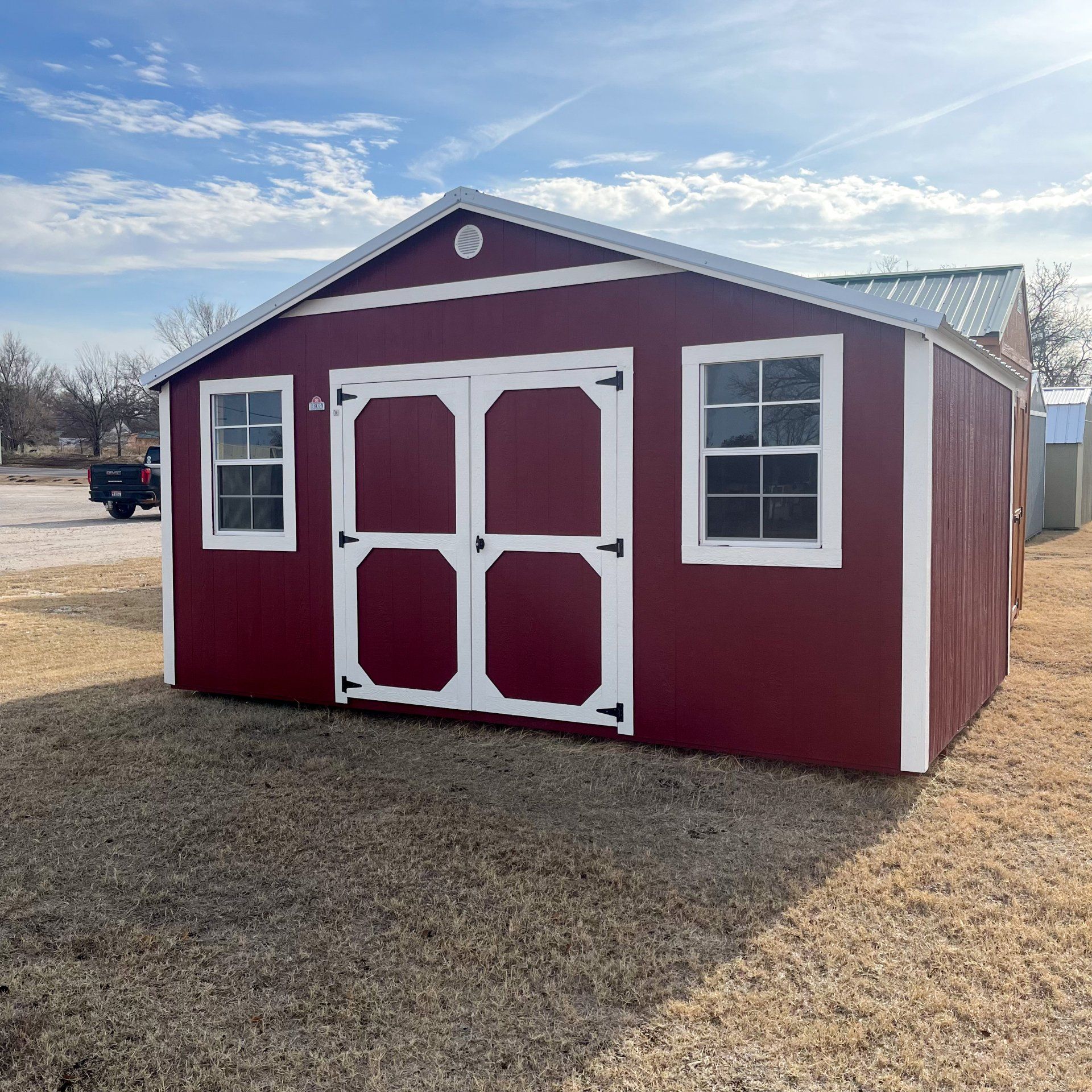 A red side utility  shed with white trim and windows is sitting in the middle of a field.