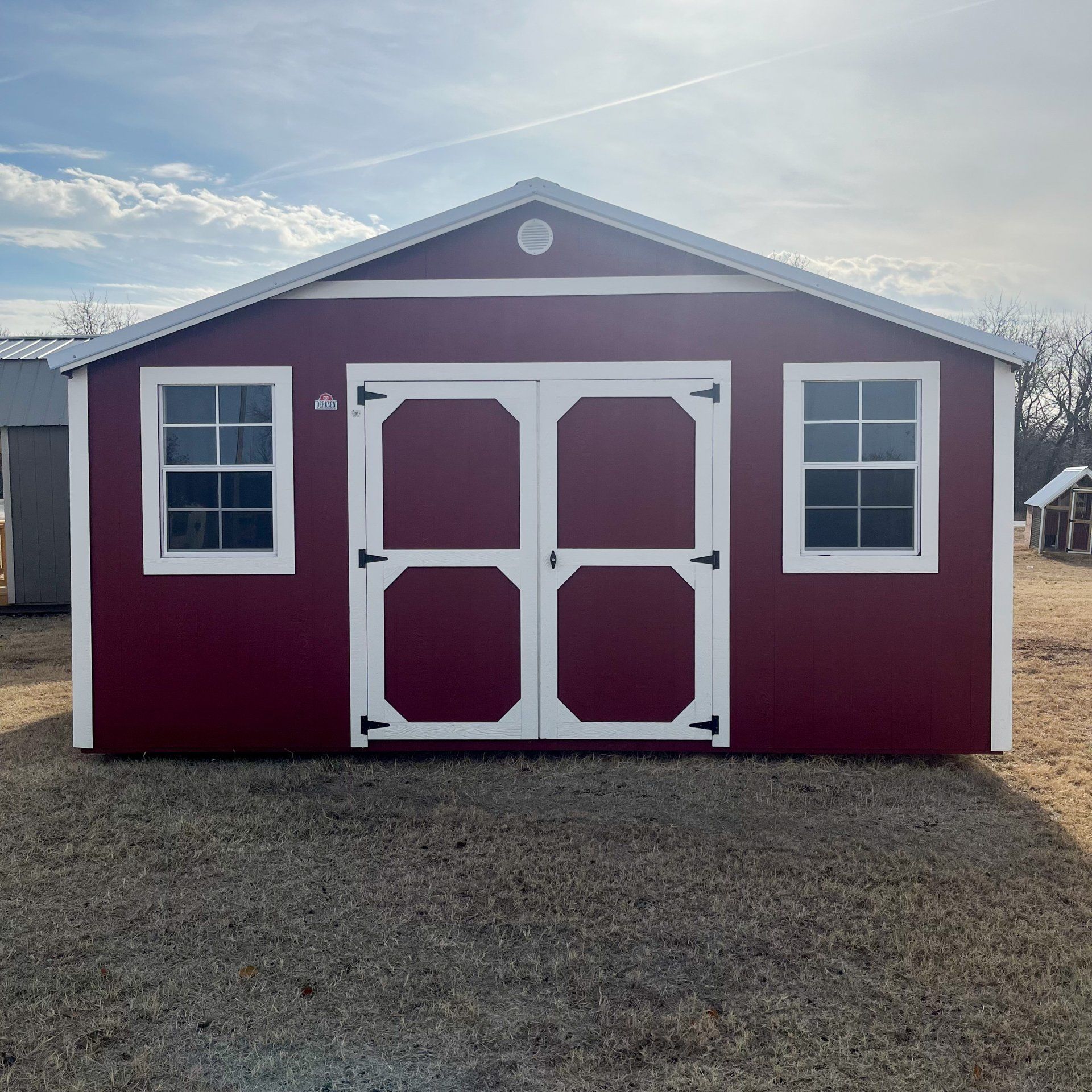 A red side utility  shed with white trim and windows is sitting on top of a dirt field.