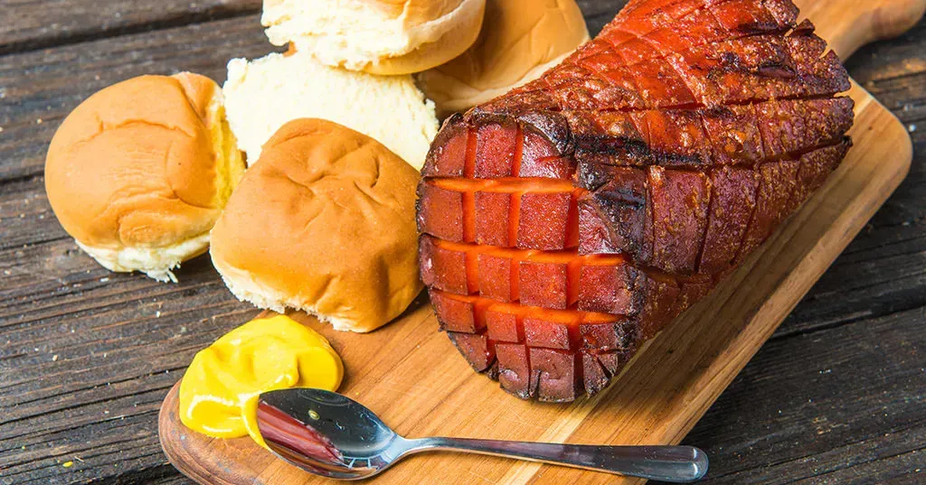 A piece of ham is sitting on a wooden cutting board next to buns and mustard.