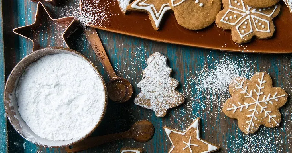 Gingerbread cookies with icing and powdered sugar, alongside cookie cutters and a bowl of sugar, on a rustic blue wooden surface.