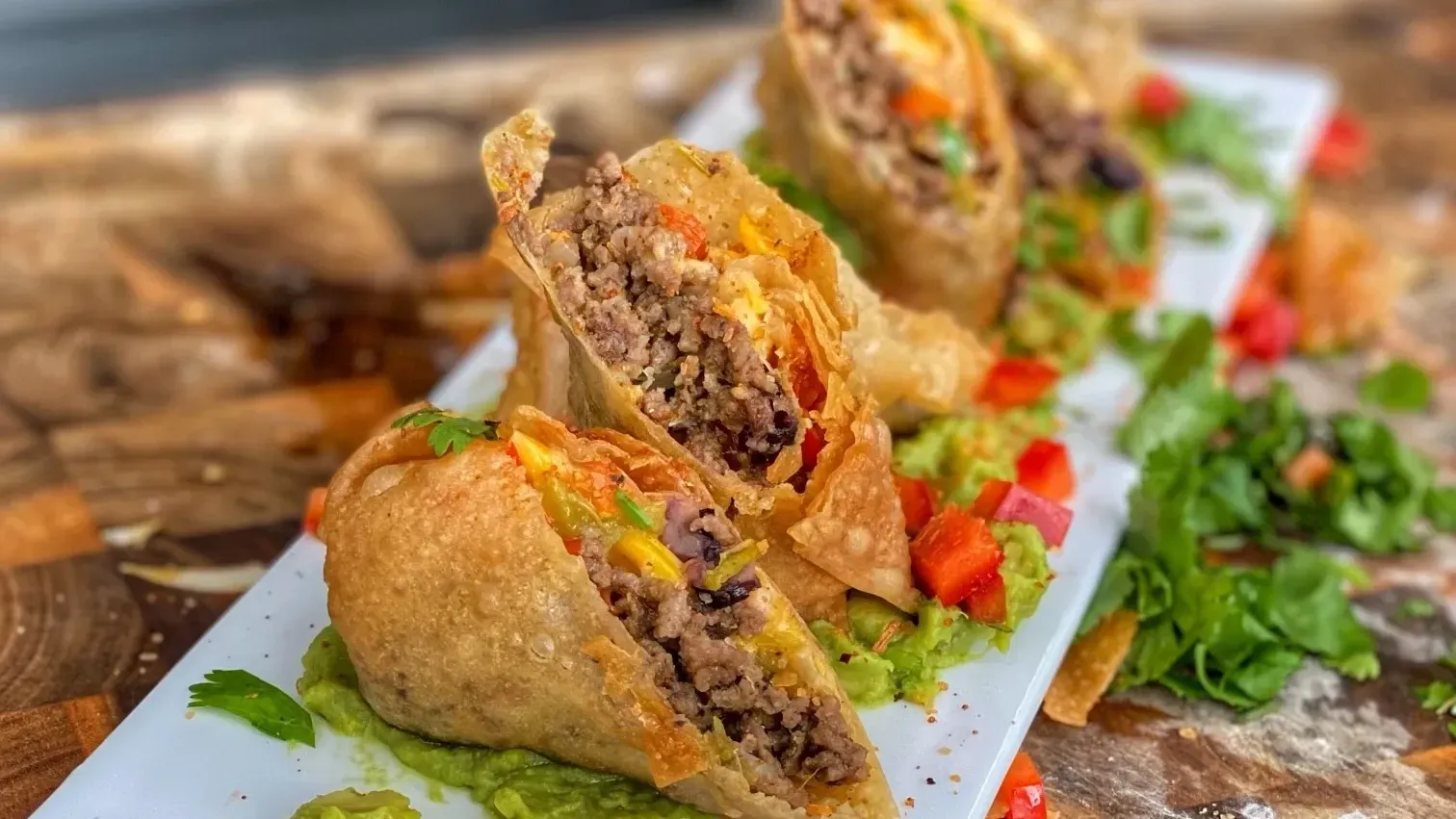 Fried burritos on a white platter with guacamole, diced vegetables, and fresh cilantro.