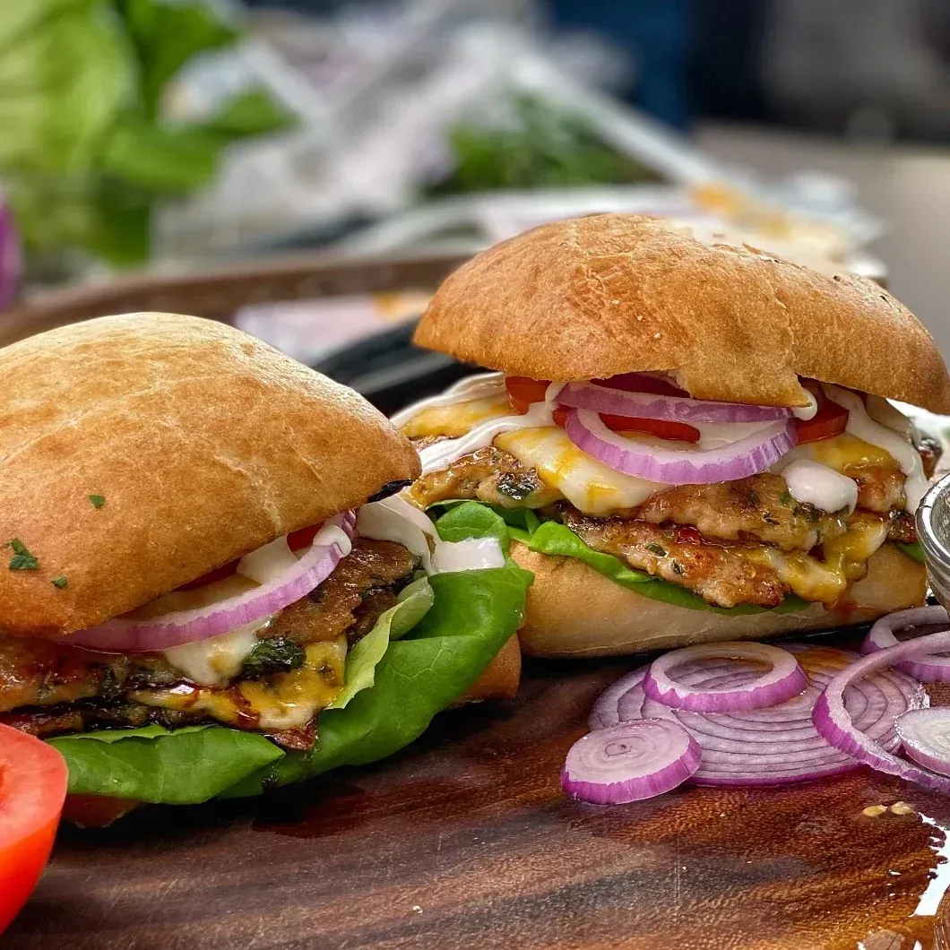 Two hamburgers with lettuce , cheese , onions and tomatoes on a wooden cutting board.