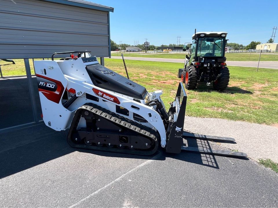 A bobcat track loader with a forklift attached to it is parked in front of a tractor.