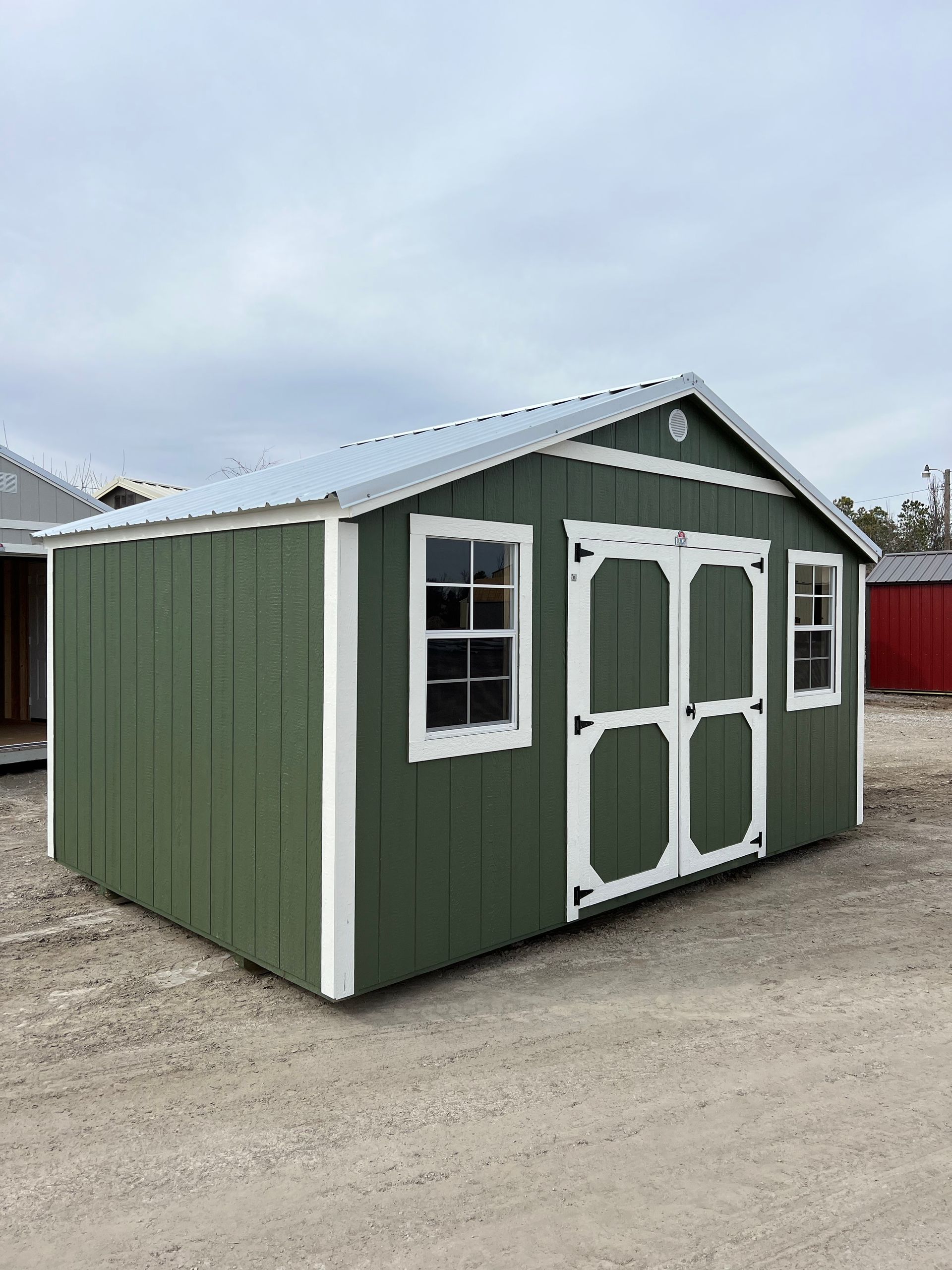 A green shed with white trim and windows is sitting on top of a dirt field.