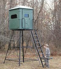 A man is standing on a ladder next to a deer stand in the woods.