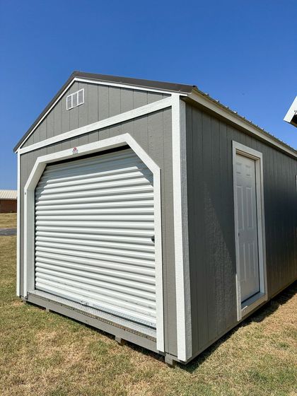 A gray garage with a white door is sitting in the middle of a grassy field.