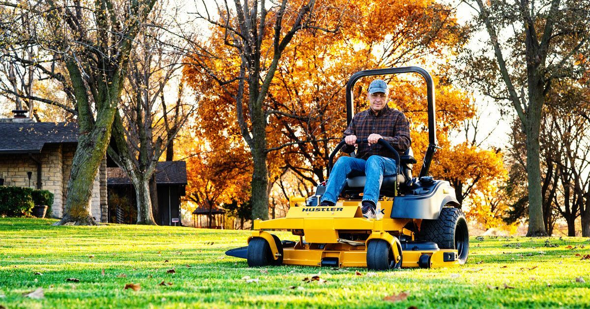 Man on ye zero-turn mower on a green lawn; fall foliage in the background