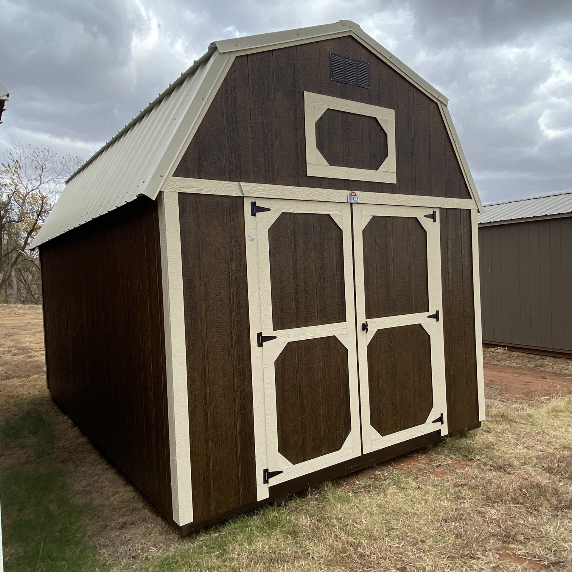 A brown barn shed with white trim is sitting in the middle of a field.