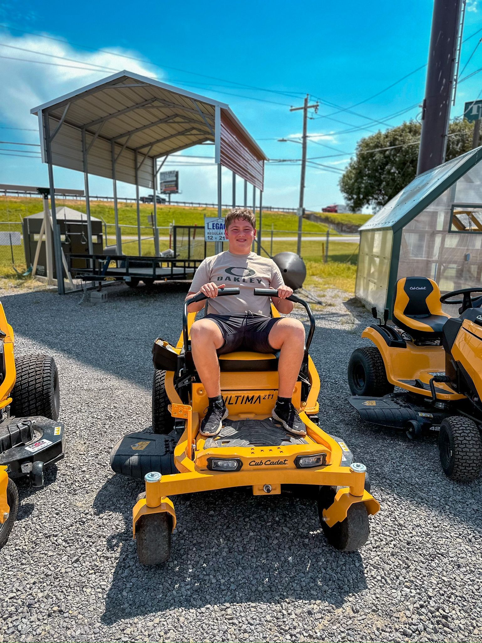Person seated on a yellow riding lawn mower in a gravel lot beside covered equipment sheds.