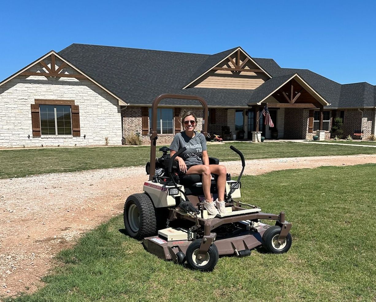 A woman is sitting on a lawn mower in front of a house.