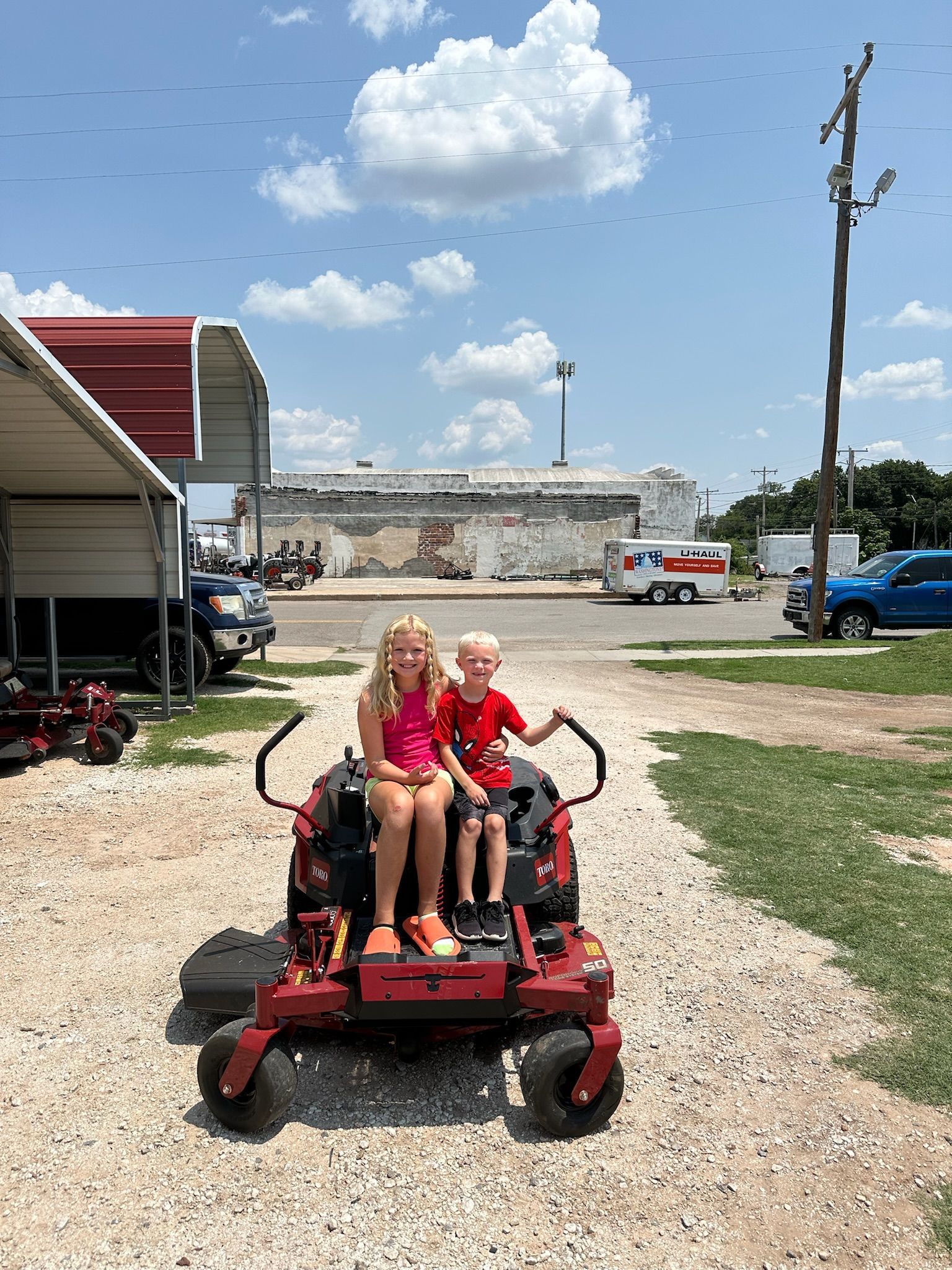 Two children are sitting on a lawn mower in a gravel lot.