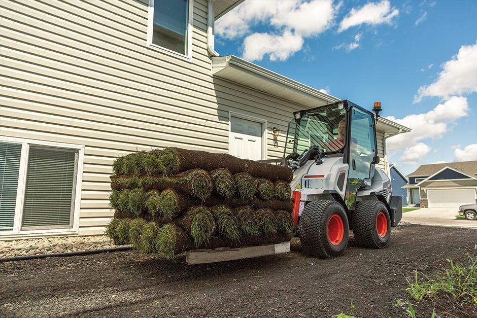 A bobcat tractor  is carrying a stack of turf in front of a house.