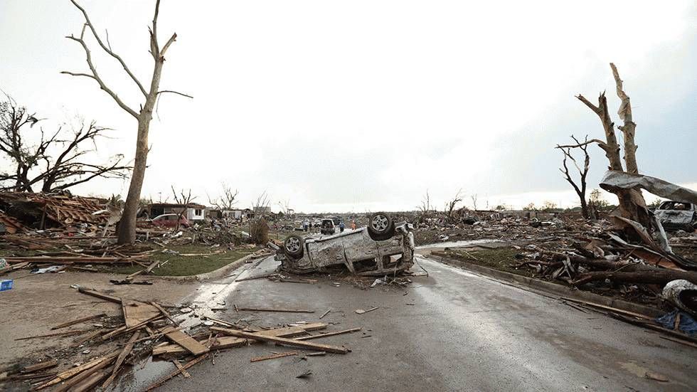 Overturned car on a debris-filled road in a tornado-ravaged area with destroyed buildings.