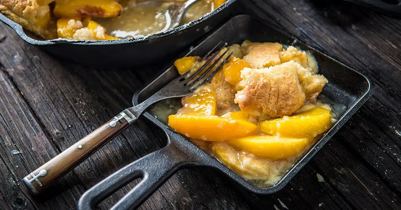 A skillet filled with peach cobbler and a fork on a wooden table.