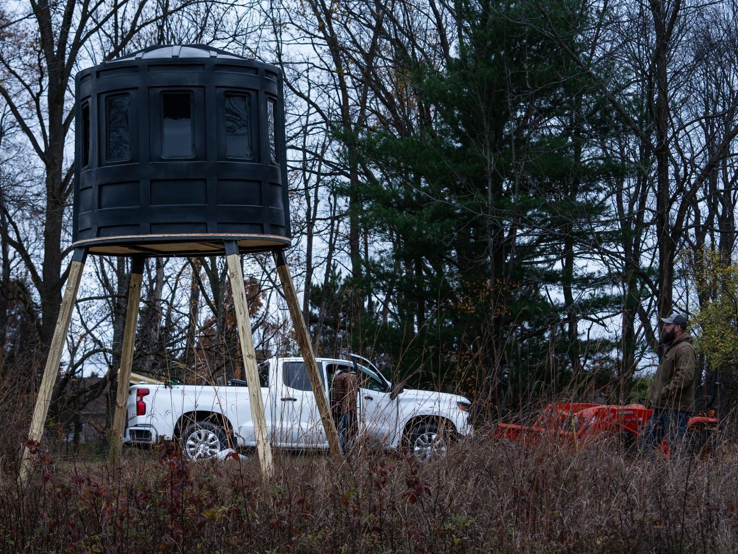 A white truck is parked next to a deer stand in the woods.