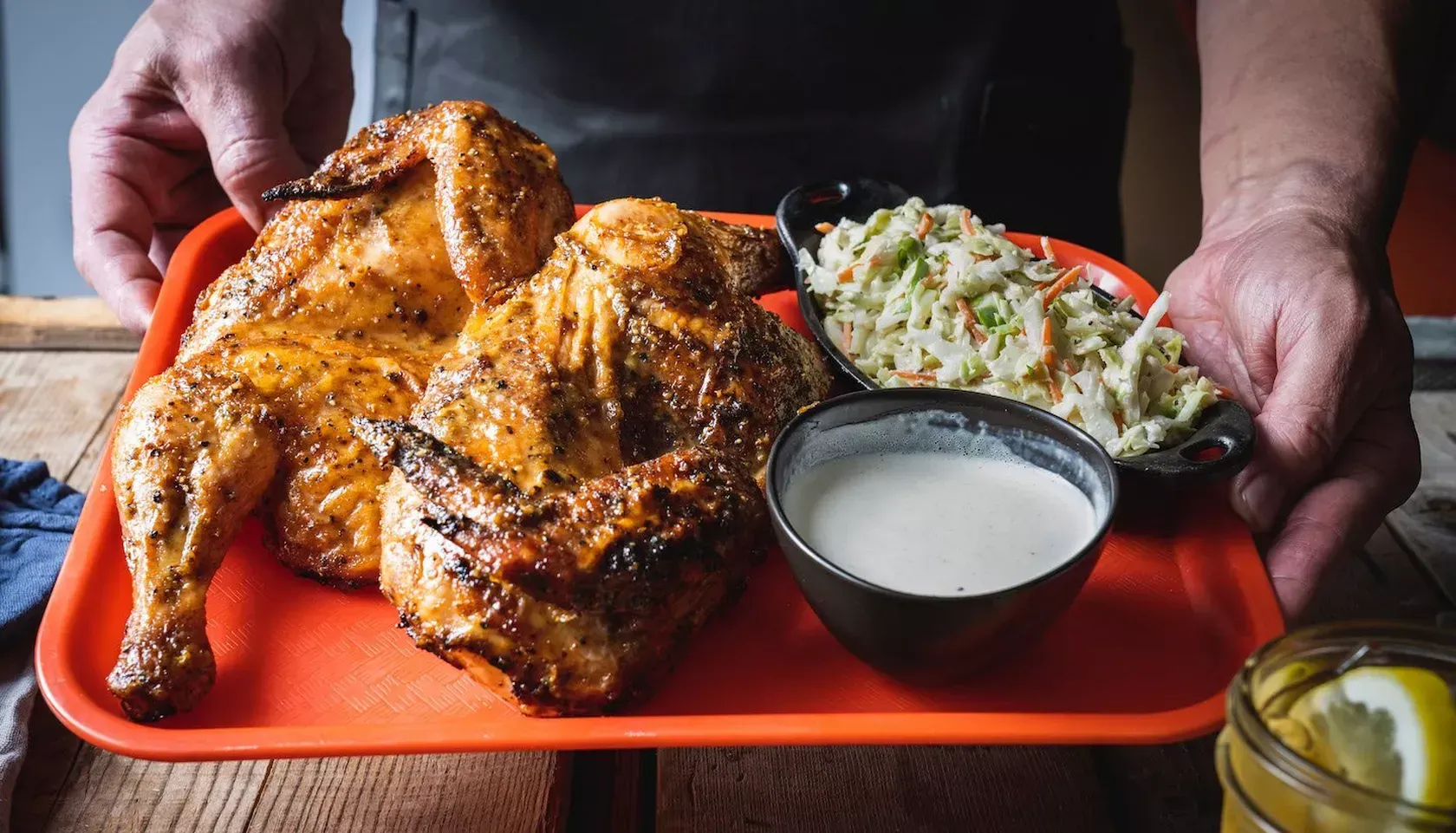 Person holding a tray with two roasted chickens, coleslaw, and white sauce.