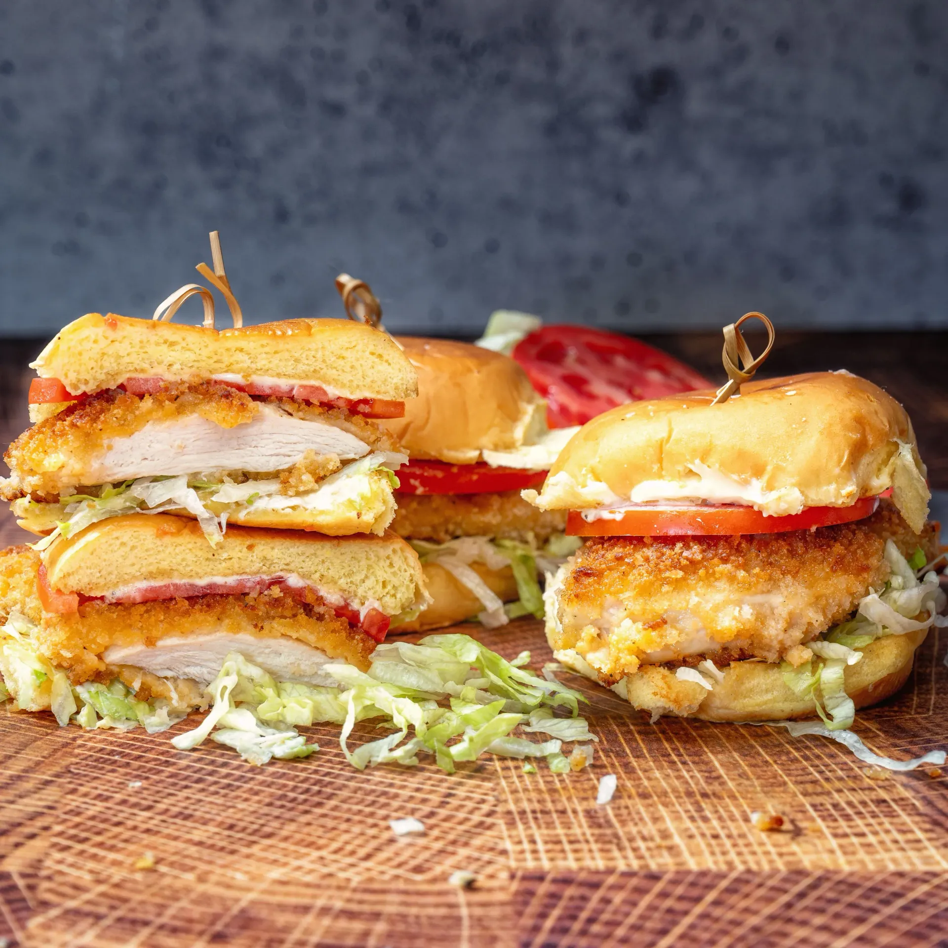 Three chicken sandwiches are stacked on top of each other on a wooden cutting board.