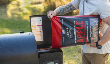 A man is holding a bag of apple wood pellets in front of a grill.
