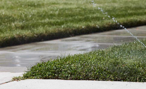 A sprinkler is spraying water on a patch of grass on the sidewalk.