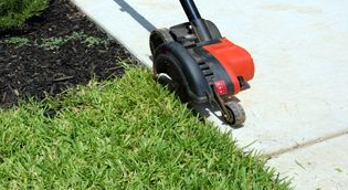 A person is using a lawn mower to cut the grass on the sidewalk.