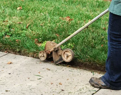 A person is using a lawn mower to cut the grass on the sidewalk.