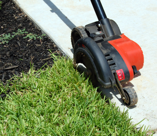 A lawn mower is cutting the grass on the sidewalk